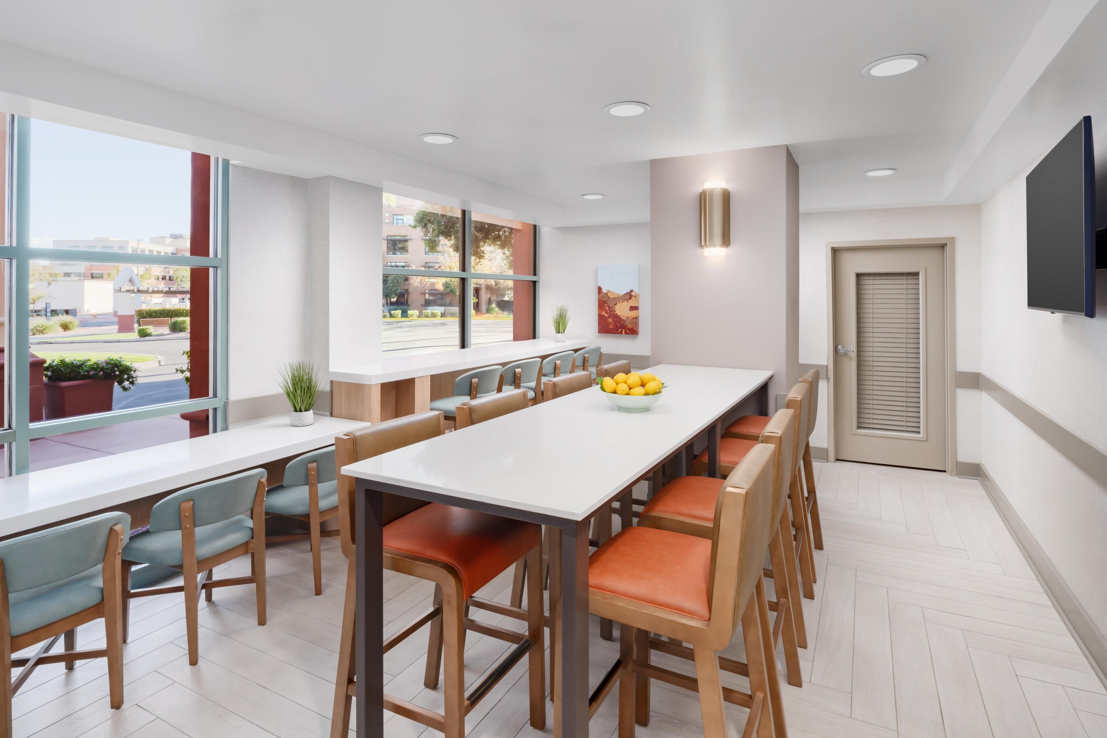 A modern kitchen with a long dining table, multiple chairs, and a large window offering a view of the outside.