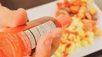 A person holding a bottle of hot sauce with a label, and a plate of food in the background.