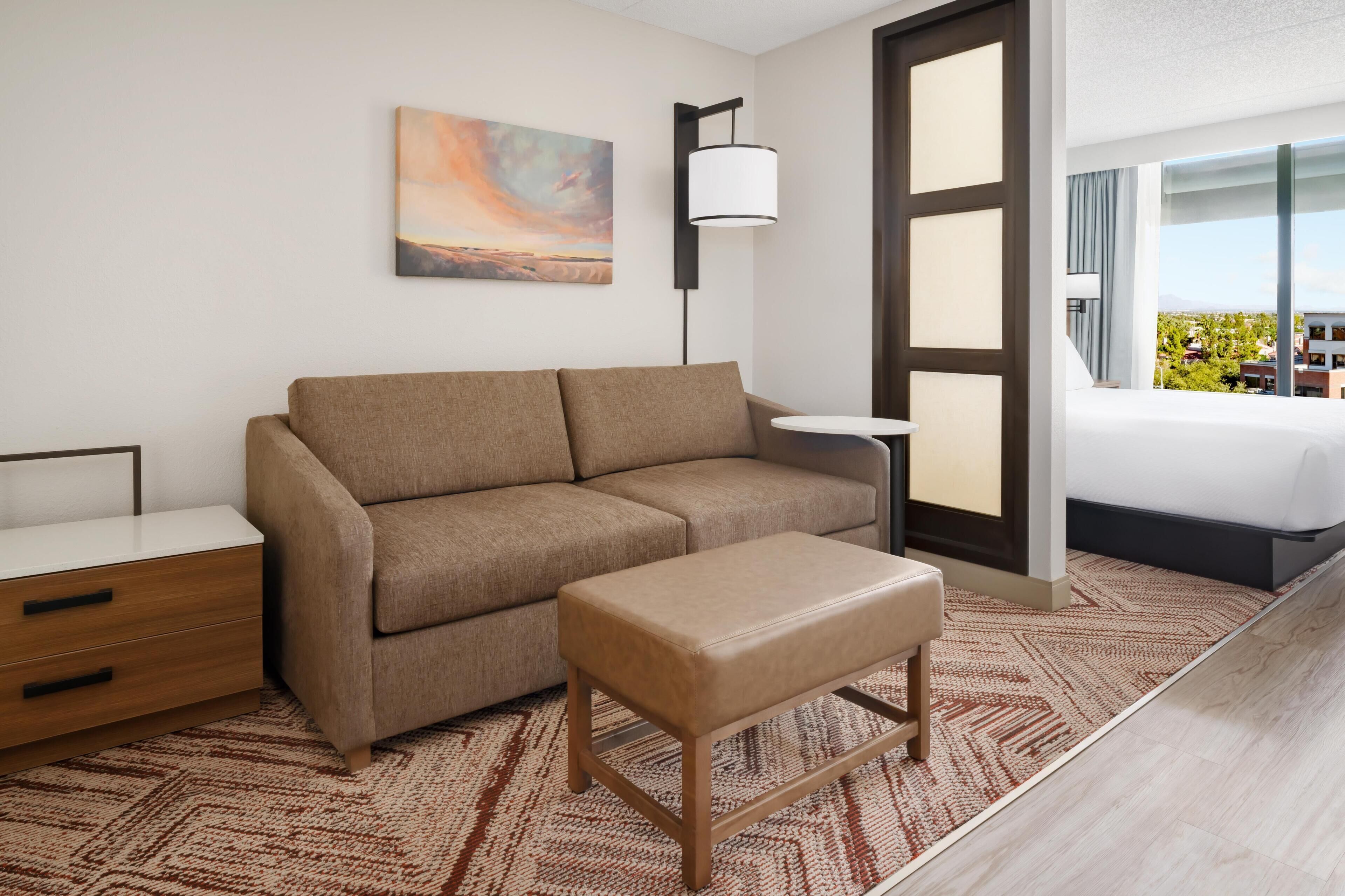 A modern hotel room with a sofa, a small table, a bench, a painting on the wall, a floor lamp, and a large window with a city view.
