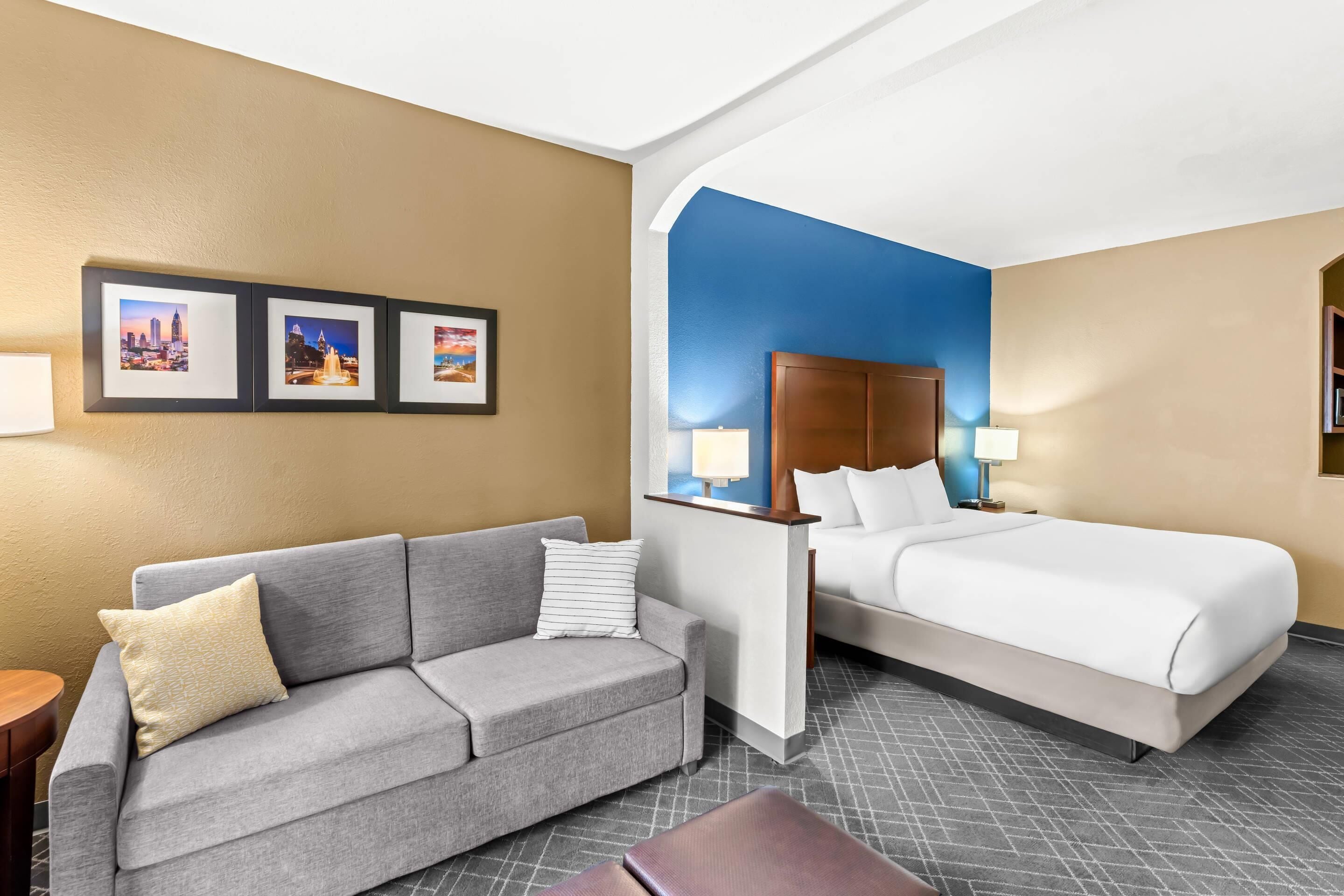 A hotel room with a grey sofa, a bed, a desk, and framed pictures on the wall.