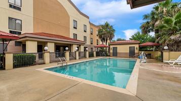 A hotel pool area with a clear blue pool, a covered seating area, and palm trees.