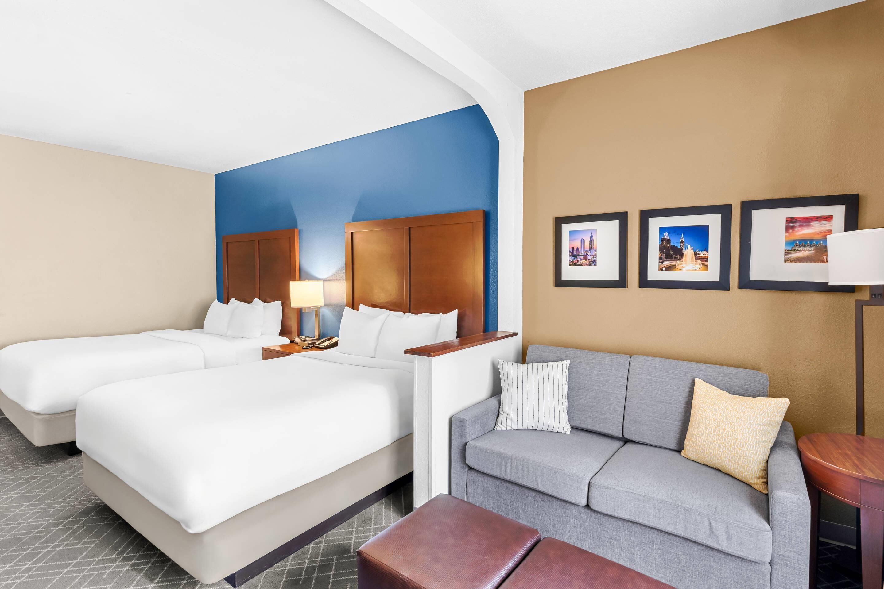 A hotel room with two beds, a gray sofa, a wooden headboard, and framed pictures on the wall.
