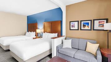 A hotel room with two beds, a gray sofa, a wooden headboard, and framed pictures on the wall.