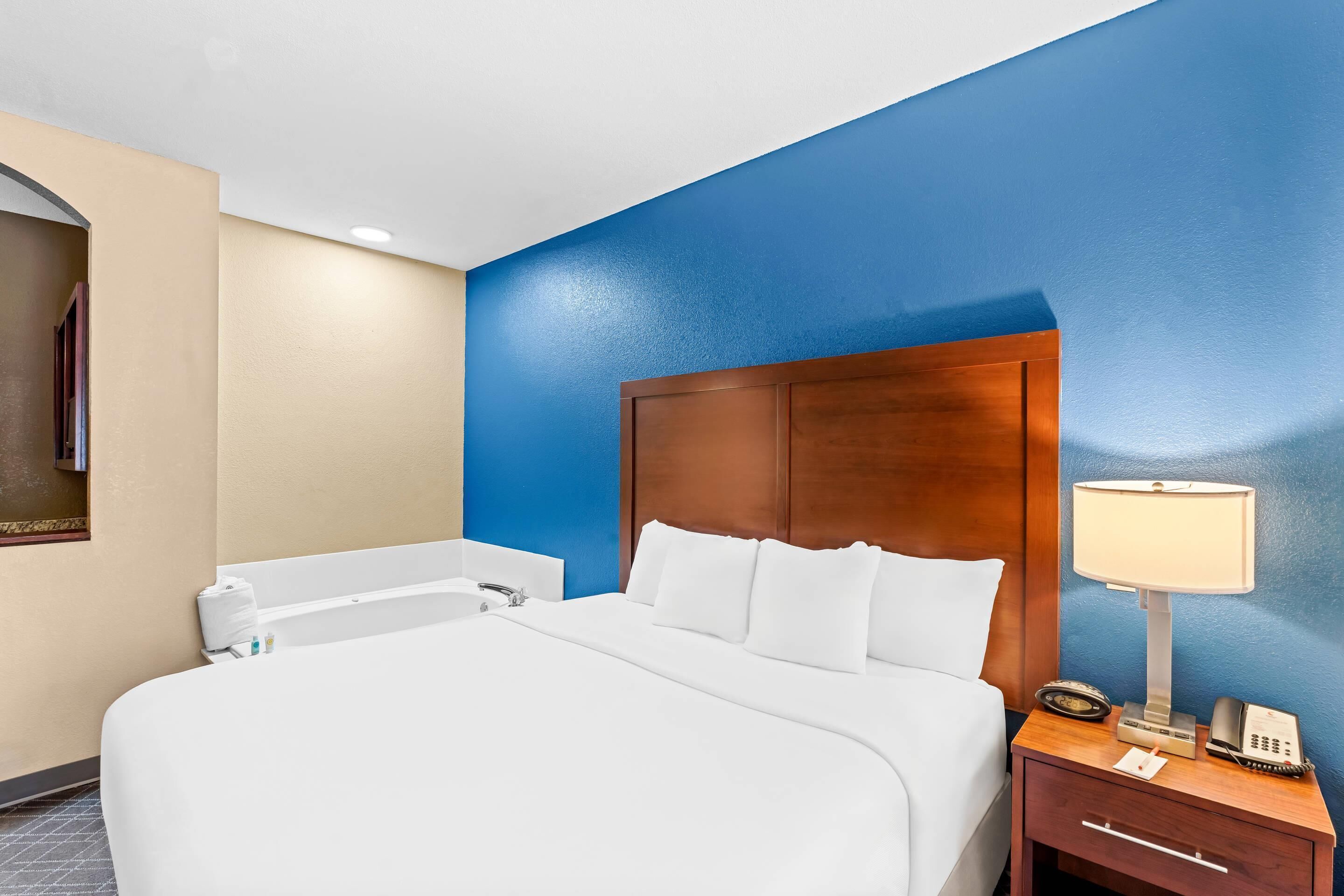 Habitación de hotel con una cama grande, cabecera de madera, mesita de noche con lámpara y baño con bañera.