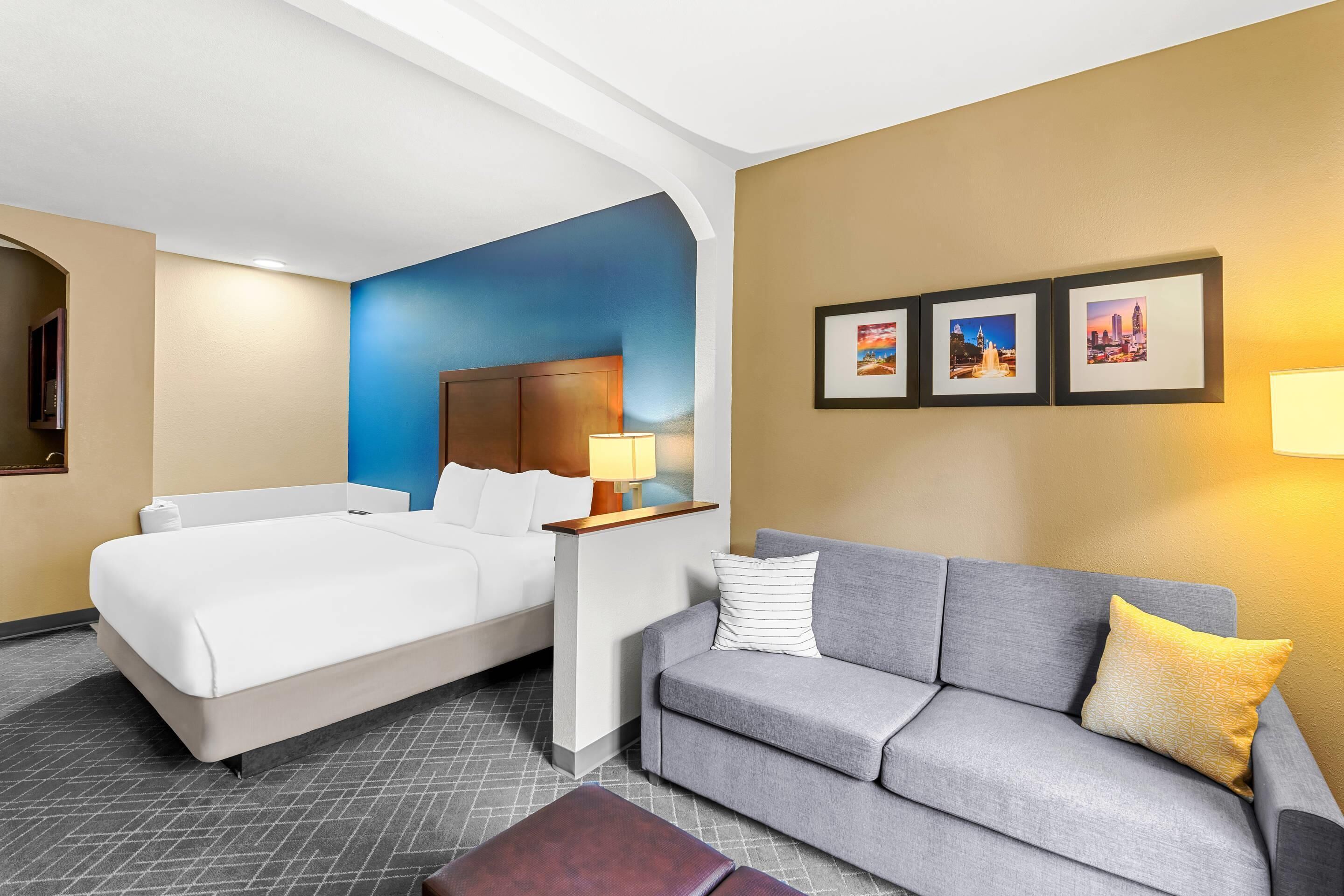 A hotel room with a bed, a grey sofa, a nightstand with a lamp, and framed pictures on the wall.