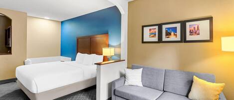 Habitación de hotel con cama, sofá gris, mesita de noche con lámpara y cuadros en la pared.