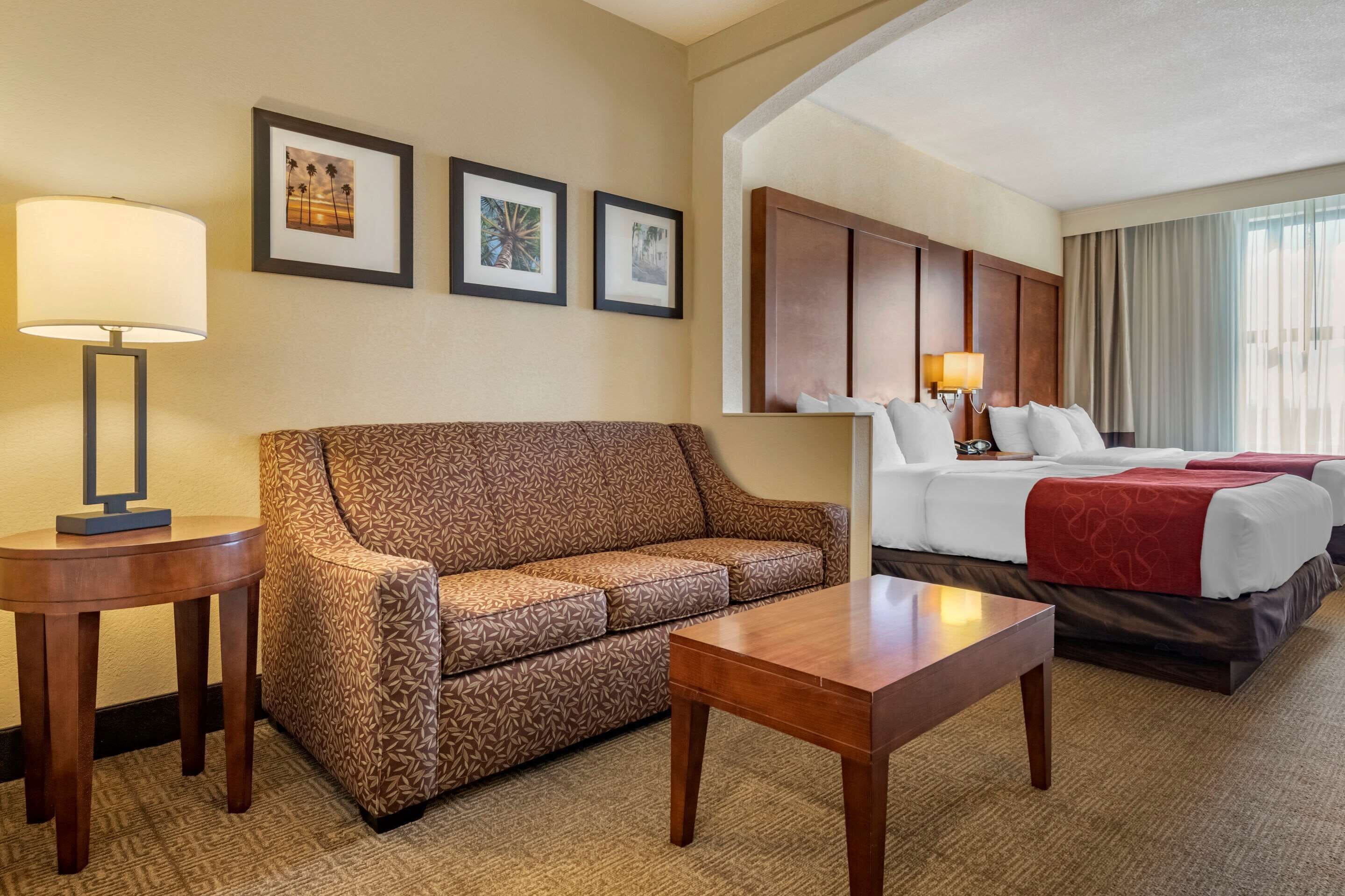 A hotel room with a patterned sofa, a wooden table, a bed with white bedding, and framed artwork on the wall.