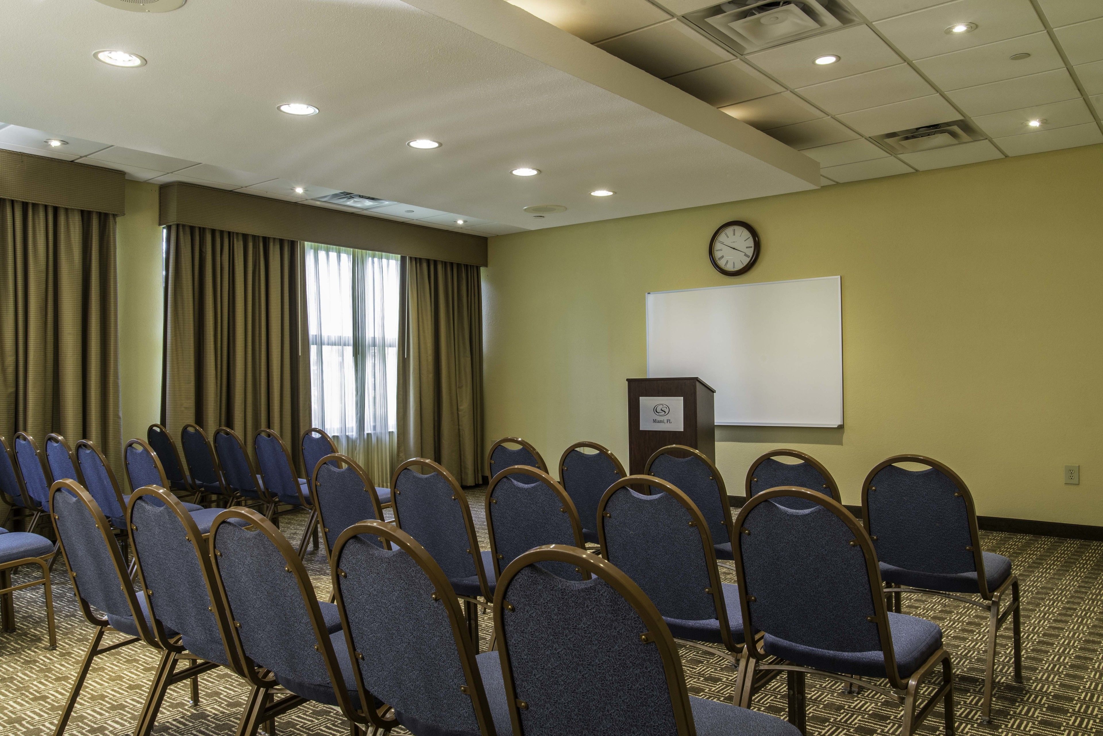 A conference room with rows of blue chairs, a whiteboard, a clock, and a podium.