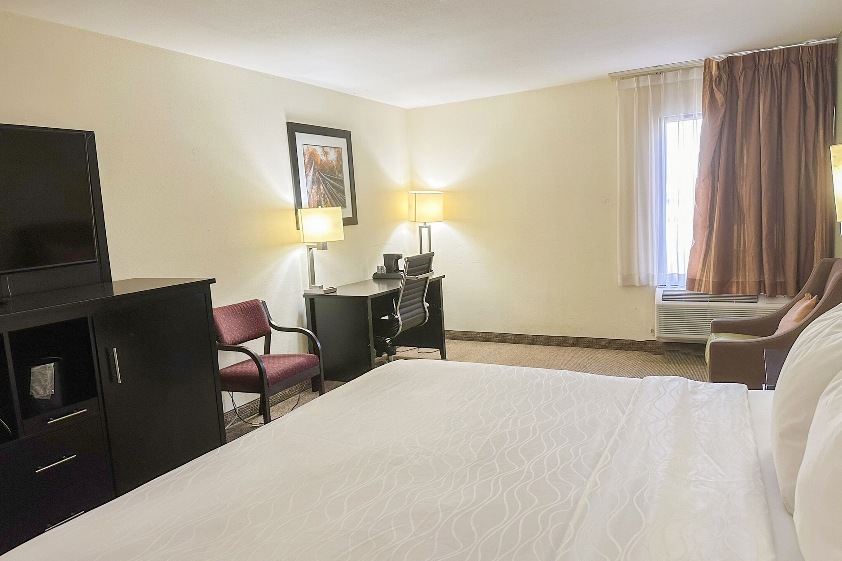 A hotel room with a large bed, a desk with a chair, a TV, and a window with curtains.