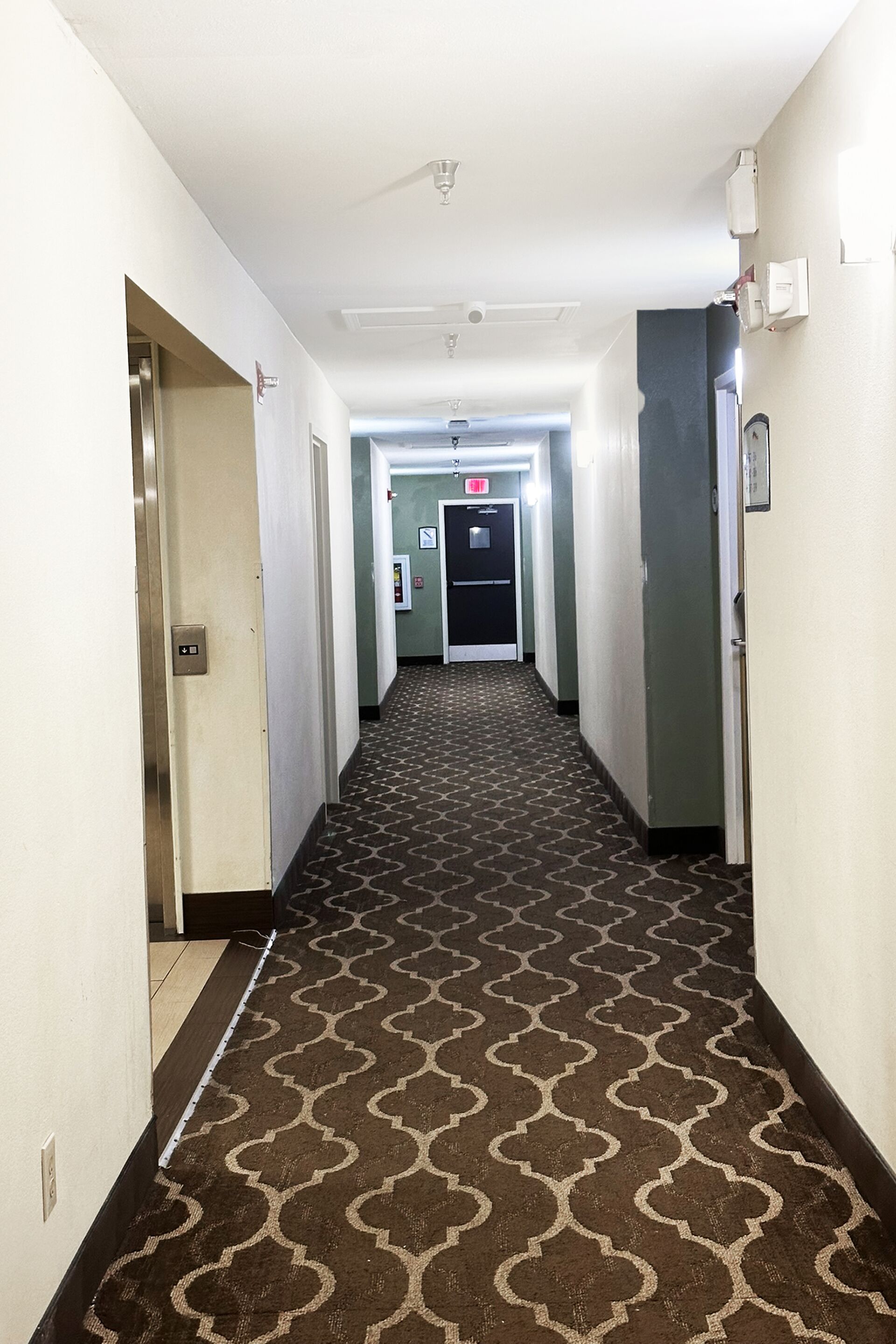 A hotel hallway with patterned carpet, a door with a light switch, and a fire alarm on the wall.