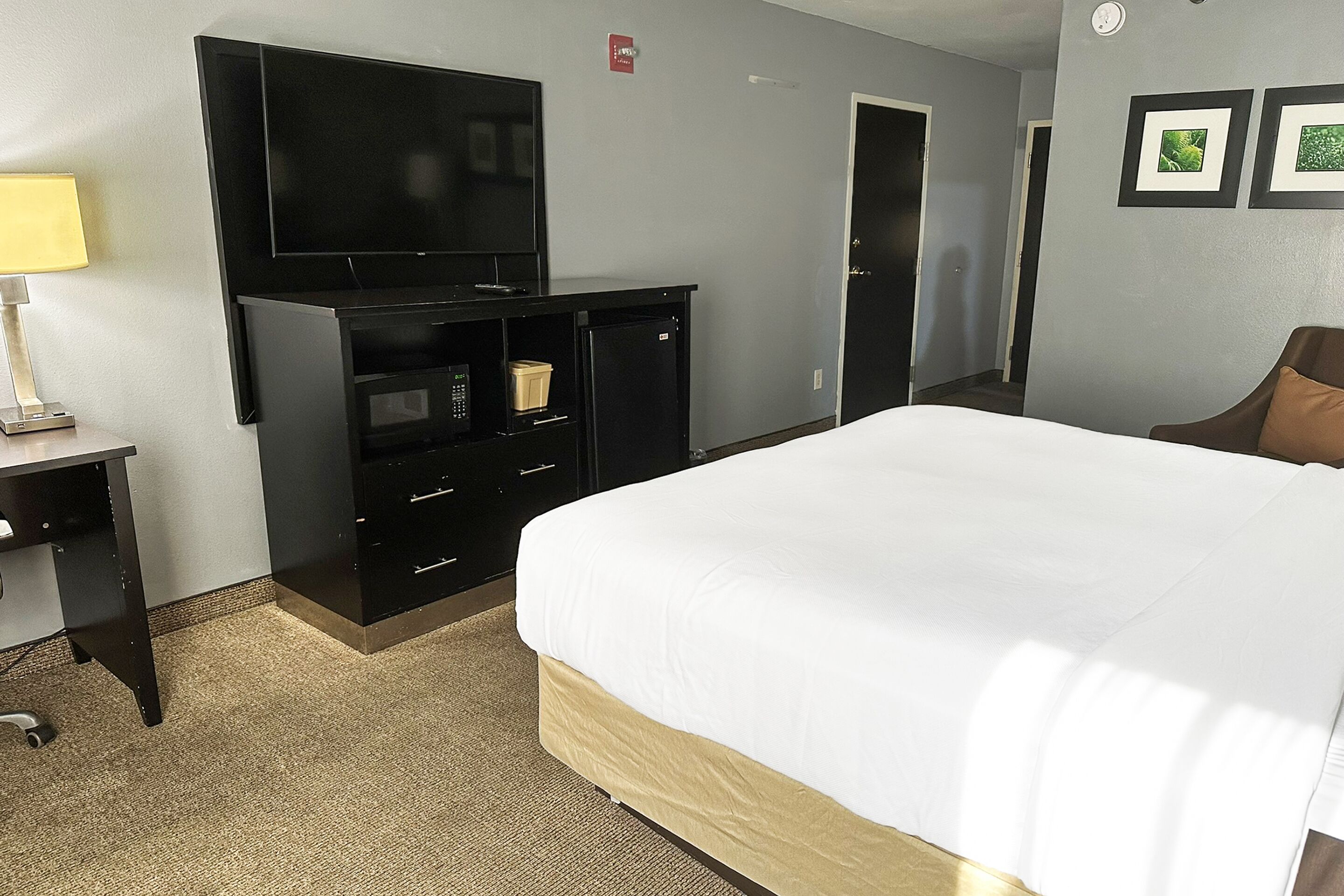 Habitación de hotel con una cama grande, un televisor de pantalla plana en un mueble negro, un escritorio con lámpara, y una puerta que da a otra habitación.
