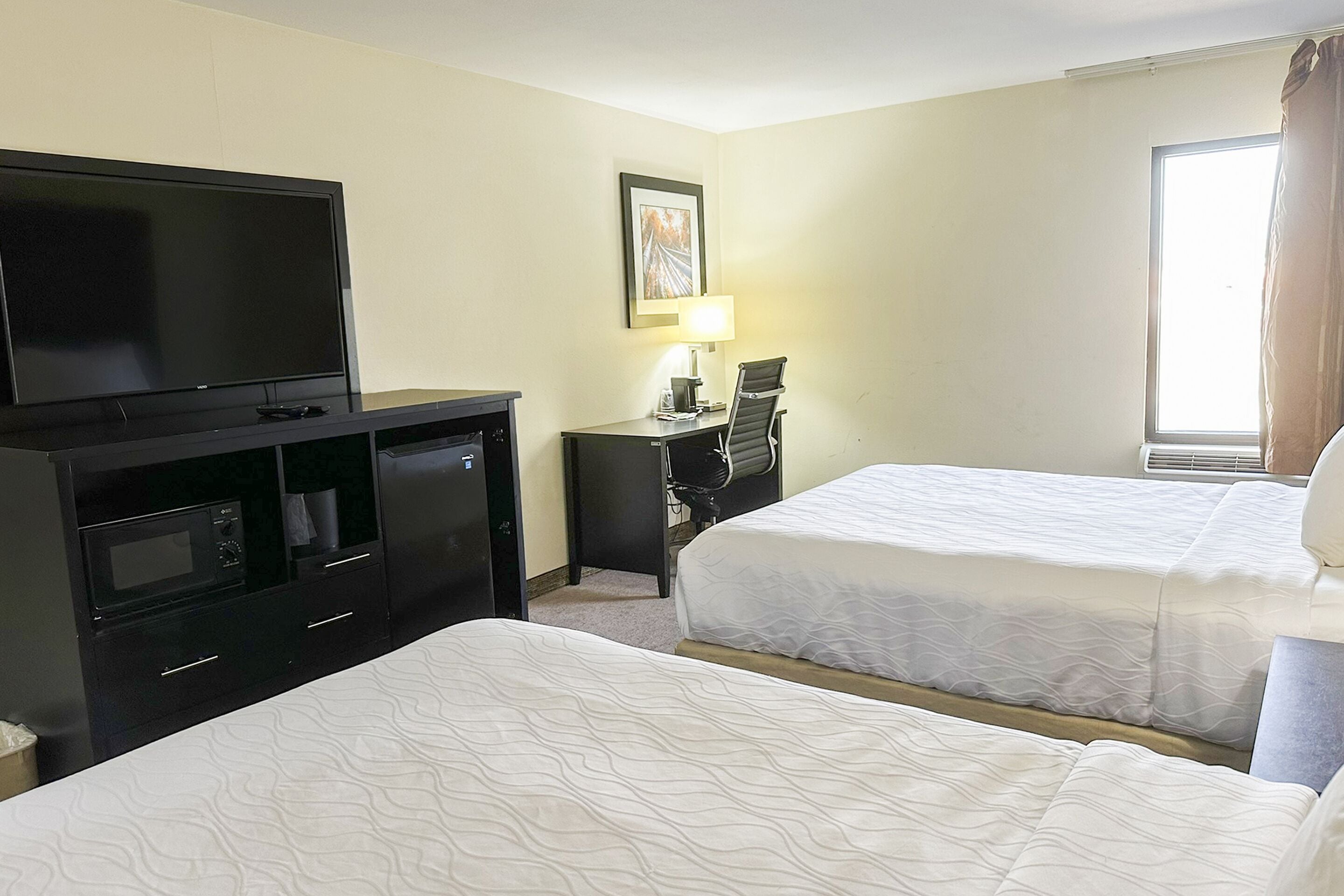 A hotel room with two beds, a TV, a desk with a lamp, and a window with curtains.