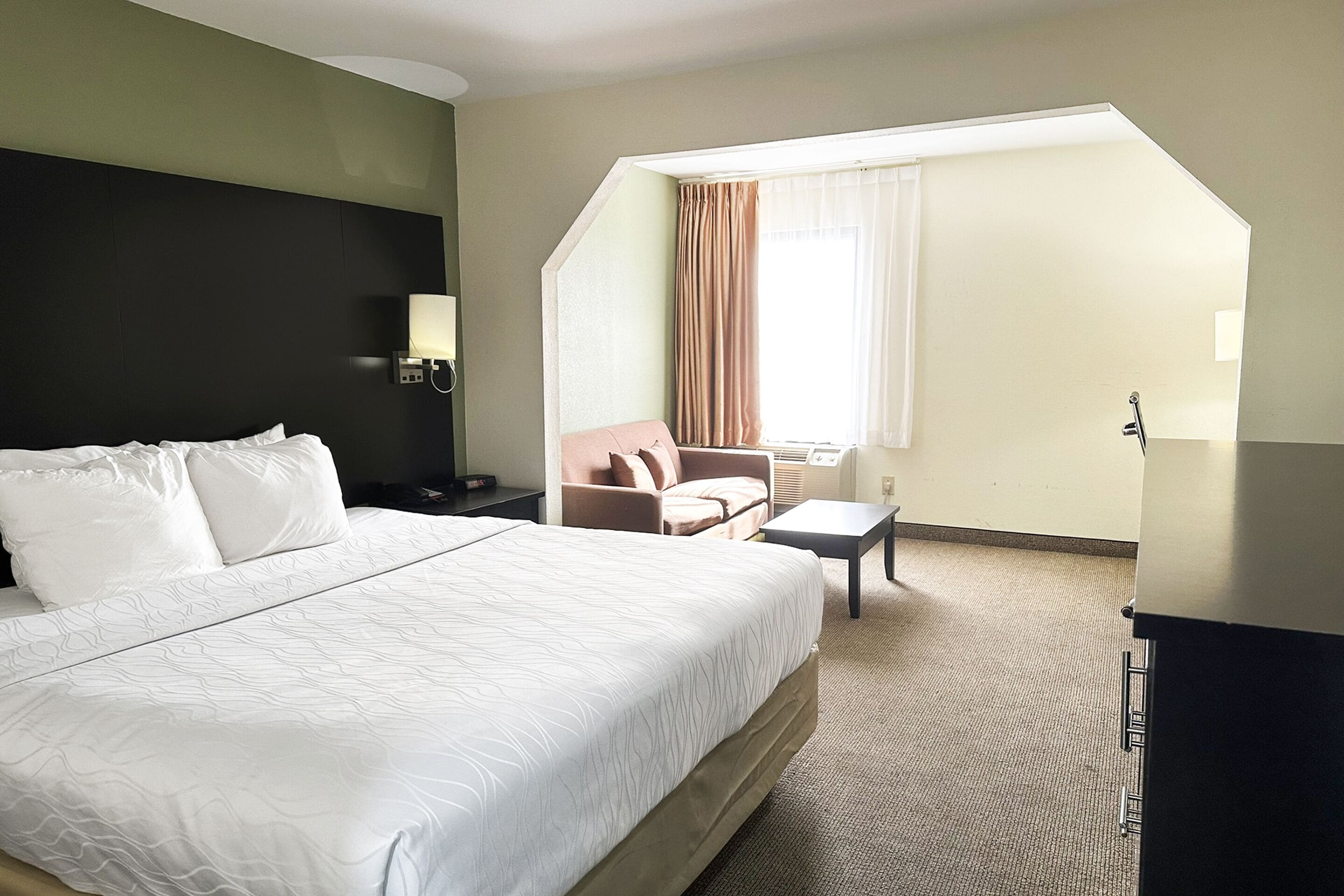 Habitación de hotel con una cama grande, un escritorio, una silla, un ventanal con cortinas y una mesita de noche con lámpara.