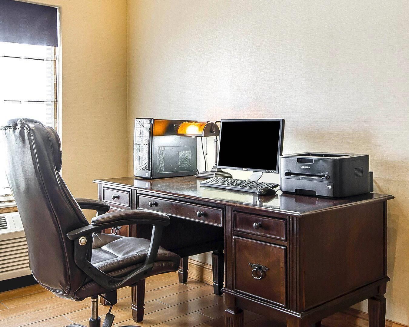 A wooden desk with a computer, printer, and chair in a room with beige walls and a window.