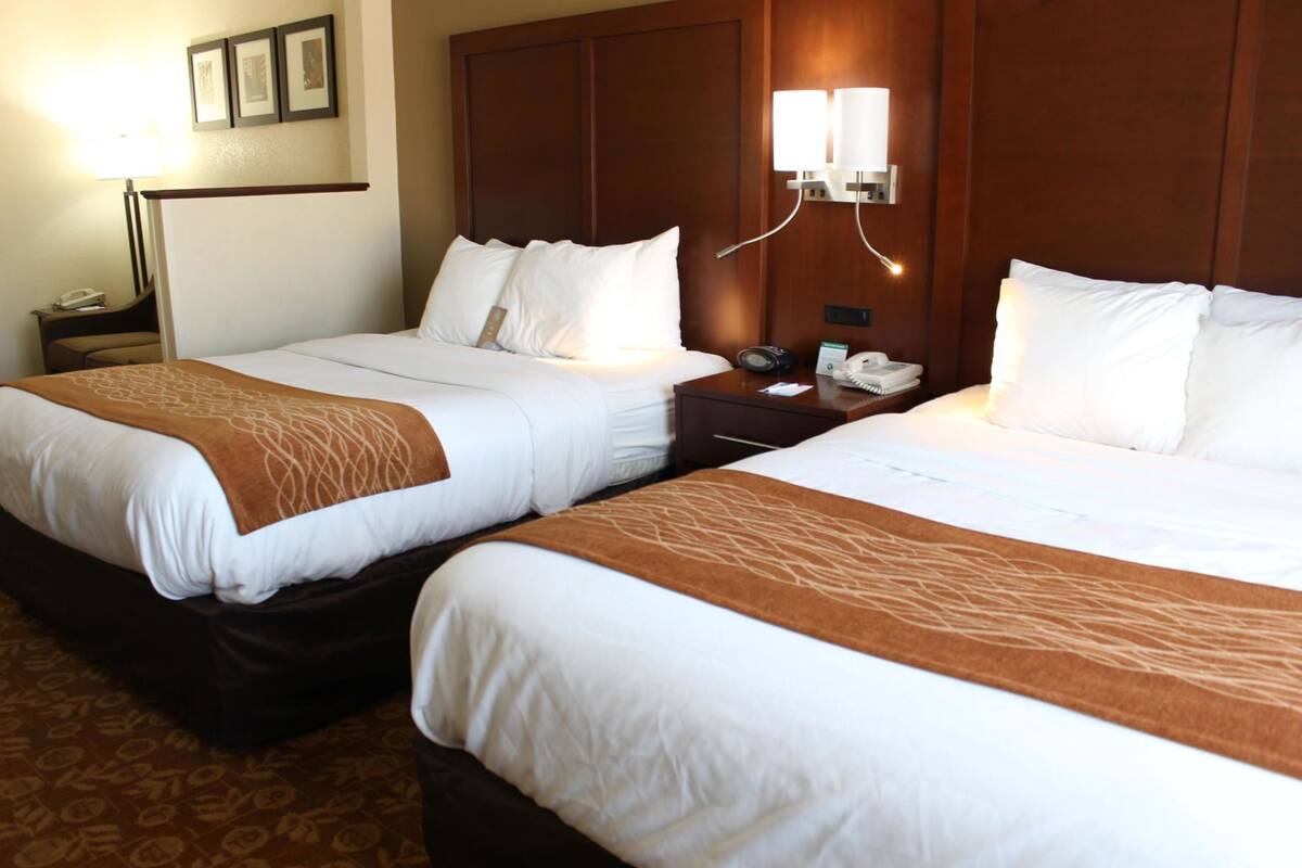 A hotel room with two beds, a nightstand with a lamp, a phone, and a wall-mounted light fixture.