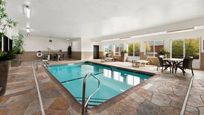 Una piscina interior con suelo de piedra, un área de descanso con sillas y una mesa, y ventanas grandes que ofrecen vistas al exterior.