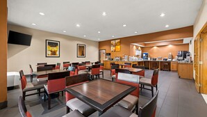 Un comedor de hotel con varias mesas y sillas, una zona de cocina y un televisor de pantalla plana montado en la pared.