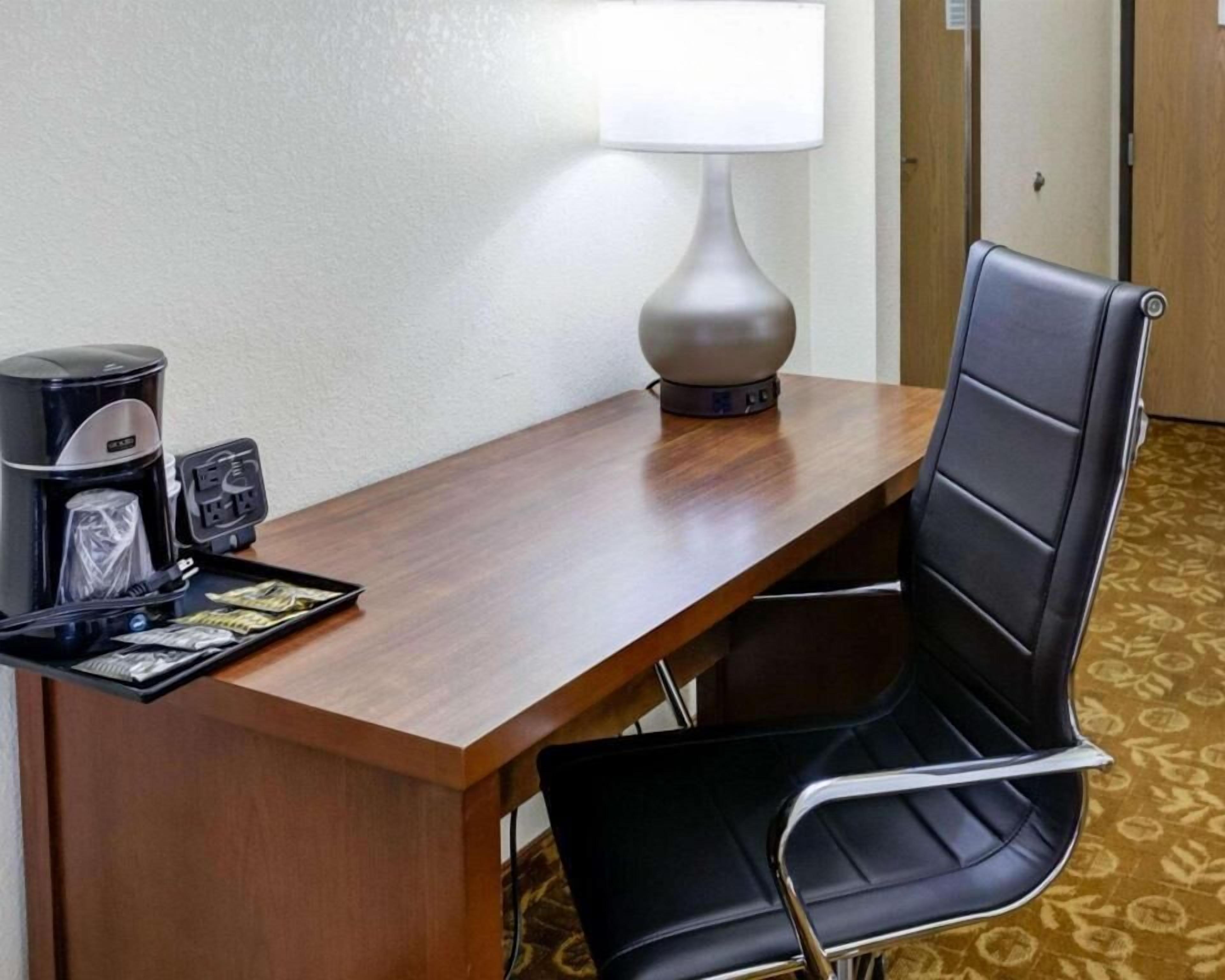 A hotel room with a wooden desk, a black chair, a lamp, a coffee maker, and a tray with a wallet and some items.