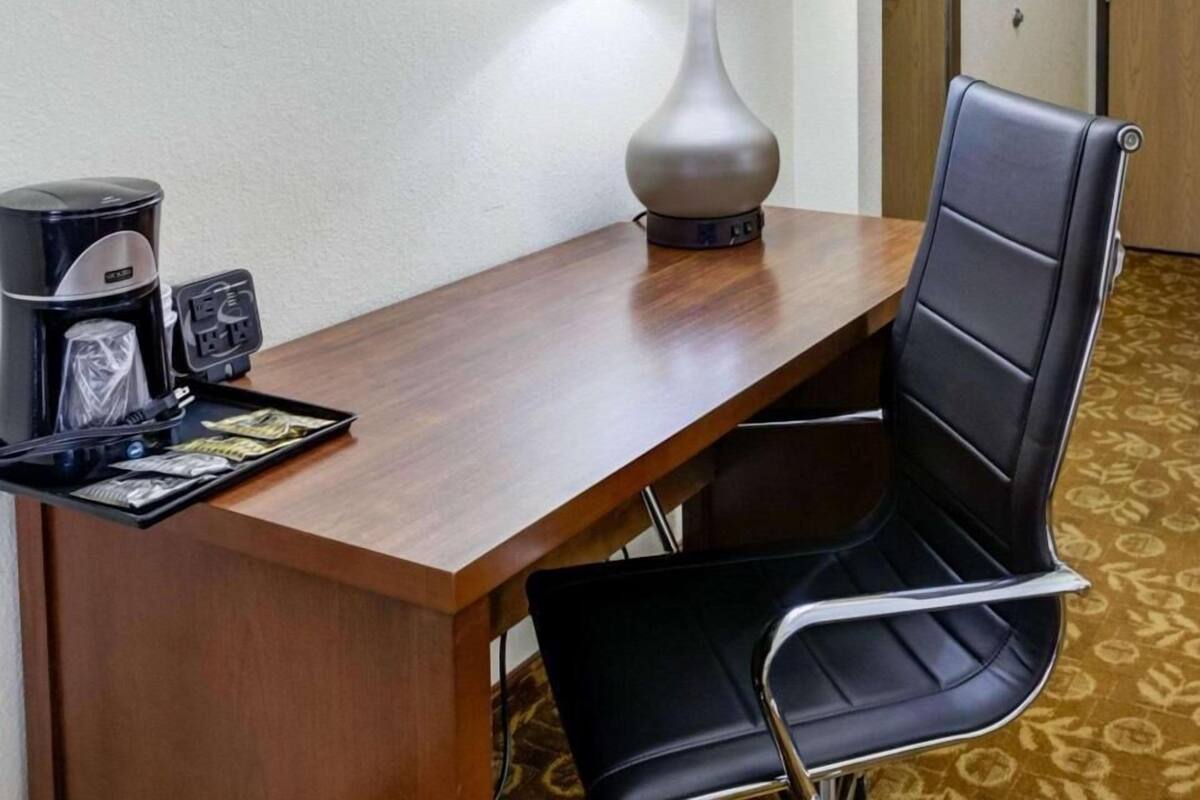 Une chambre d’hôtel avec un bureau en bois, une chaise noire, une lampe, une machine à café et un plateau contenant un portefeuille et quelques objets.
