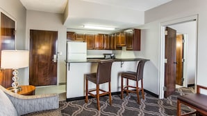 A compact kitchen with wooden cabinets, a microwave, and a small countertop. There are two wooden bar stools and a grey sofa in the living area.