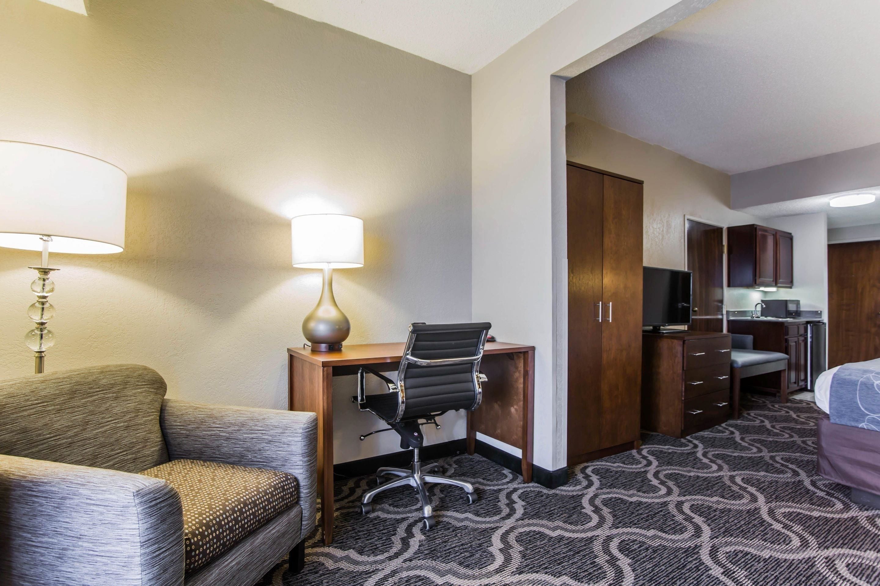 A hotel room with a sofa, a desk with a chair, a lamp, a kitchenette, and a bed.