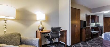 A hotel room with a sofa, a desk with a chair, a lamp, a kitchenette, and a bed.