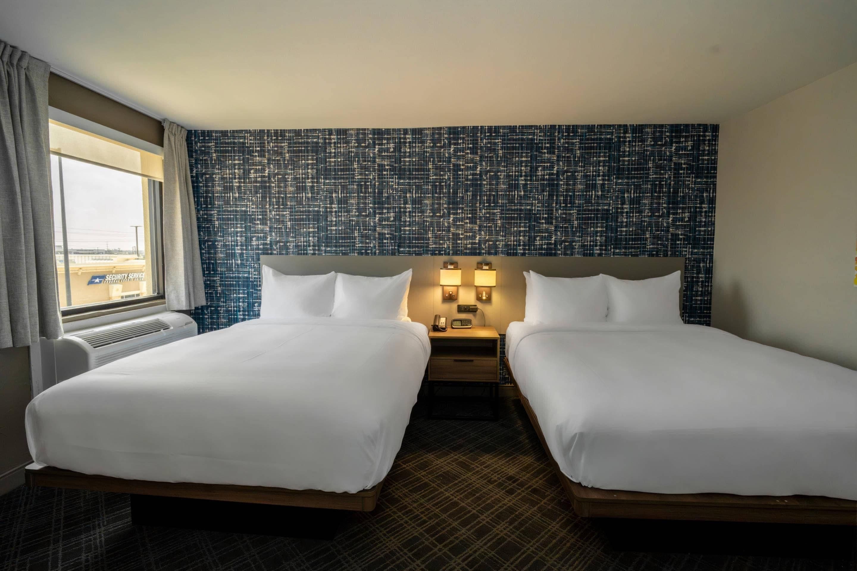 A hotel room with two beds, a window with curtains, a wall with a textured pattern, and a nightstand with a lamp.