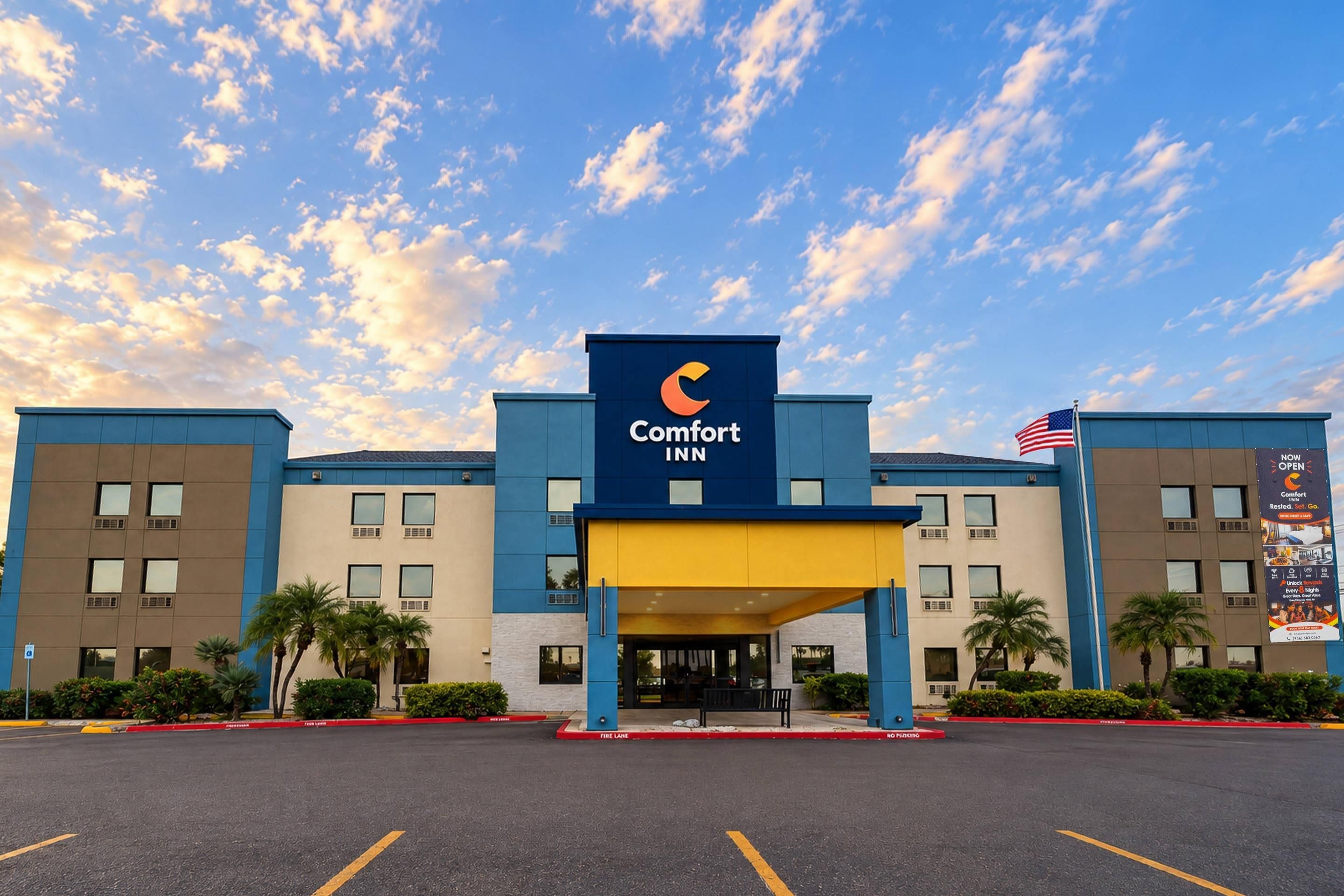 Un hotel Comfort Inn con fachada azul y amarilla, una bandera estadounidense y un cielo despejado con nubes.