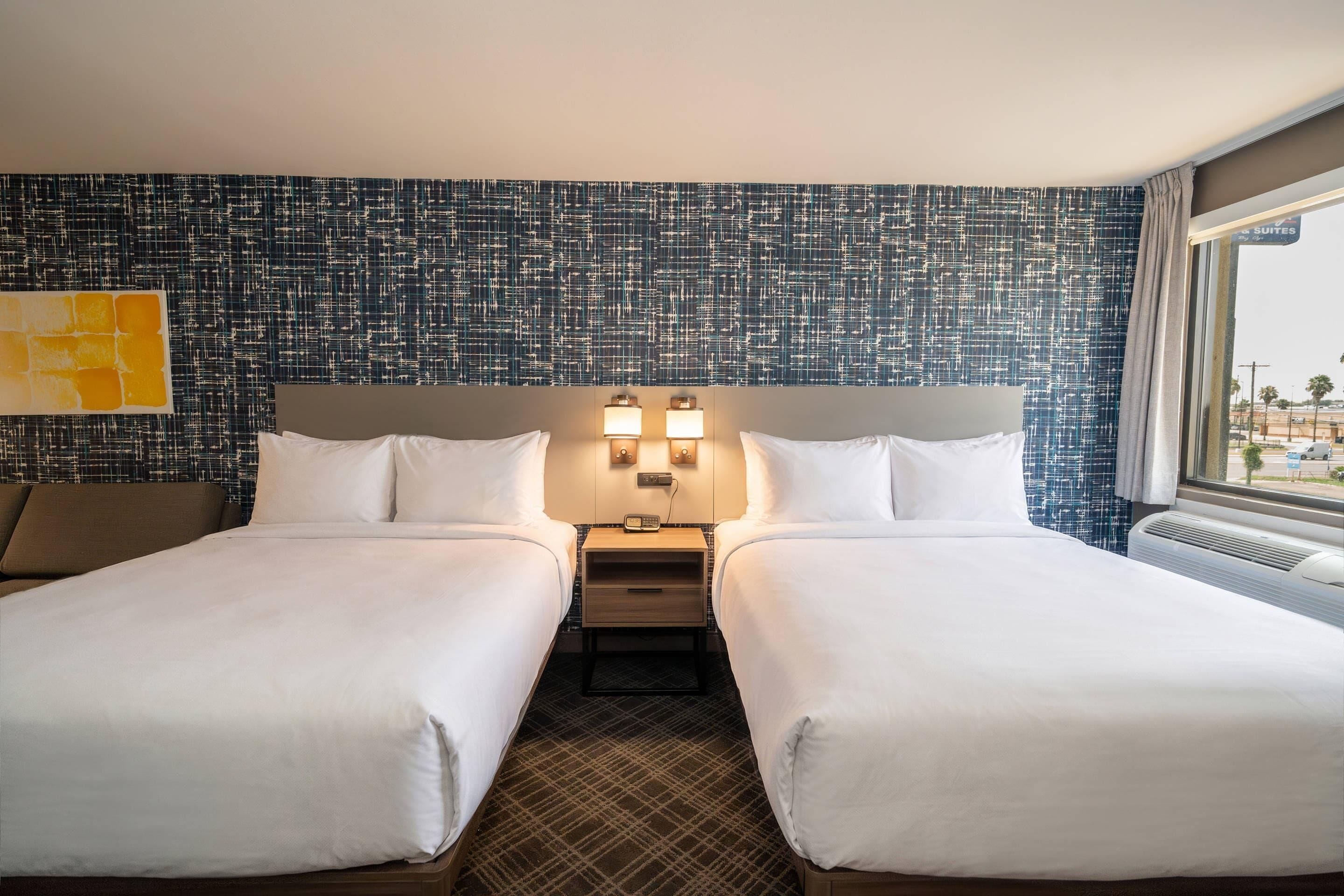 A hotel room with two beds, a nightstand with a lamp, a large window, and a patterned wall.