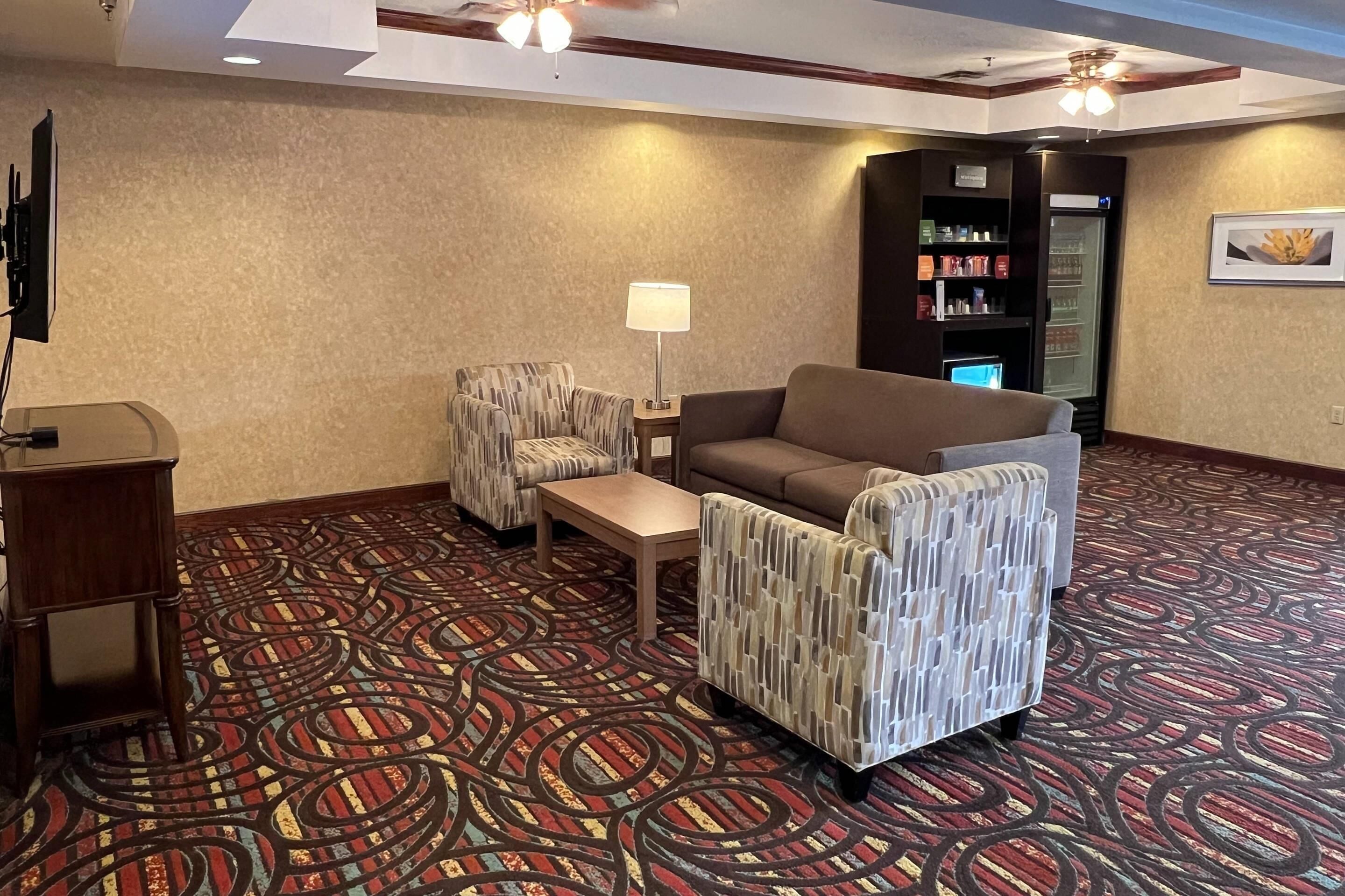Un hall d’hôtel avec un tapis à motifs, un canapé, deux fauteuils, une petite table, une lampe, un téléviseur, un réfrigérateur et un distributeur automatique.
