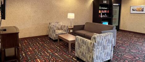 Un hall d’hôtel avec un tapis à motifs, un canapé, deux fauteuils, une petite table, une lampe, un téléviseur, un réfrigérateur et un distributeur automatique.