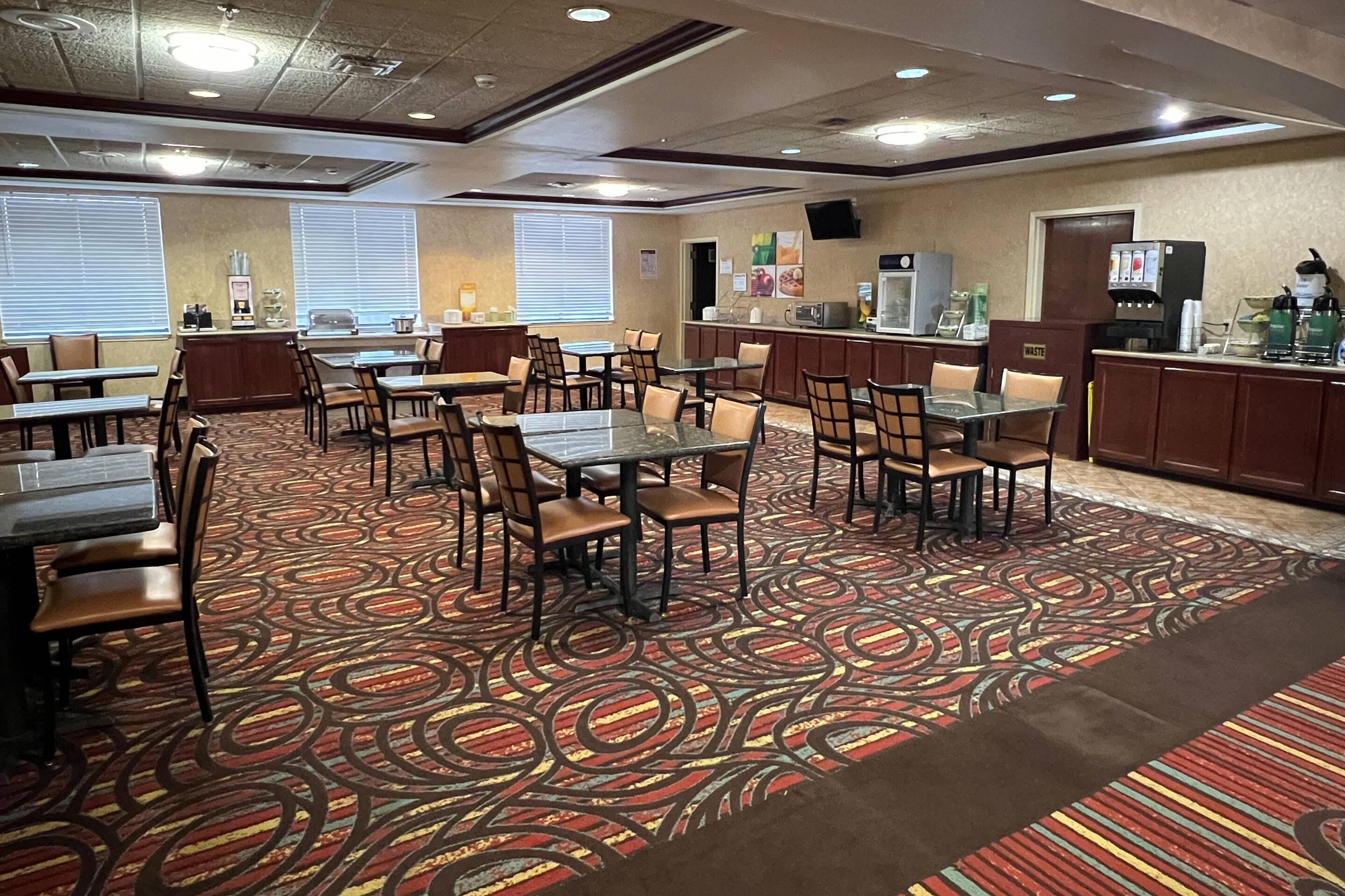 A hotel dining area with multiple tables and chairs, a kitchen counter, and a waste bin.