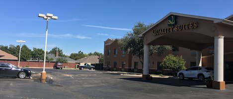 A hotel entrance with a sign reading "Quality Suites," a parking lot with several cars, and a clear blue sky.