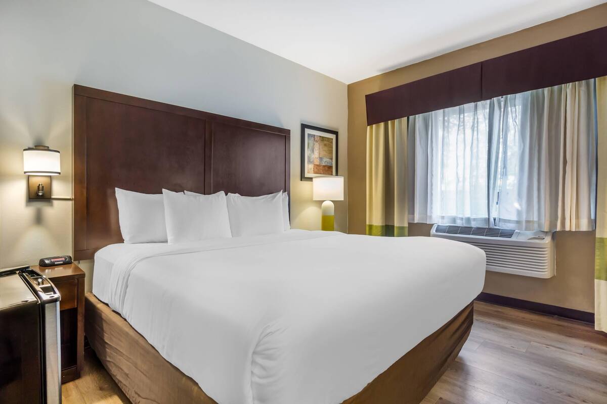 A large bed with white linens, a wooden headboard, and a nightstand with a lamp. There is a window with curtains and an air conditioning unit on the wall.