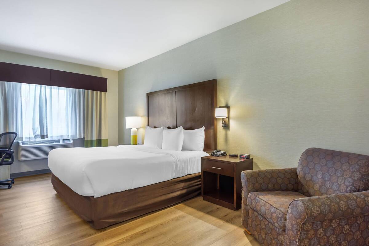 A hotel room with a bed, a nightstand, a lamp, a chair, and a window with curtains.