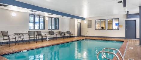 Una piscina interior con agua cristalina, rodeada de sillas y mesas, y un suelo de baldosas.