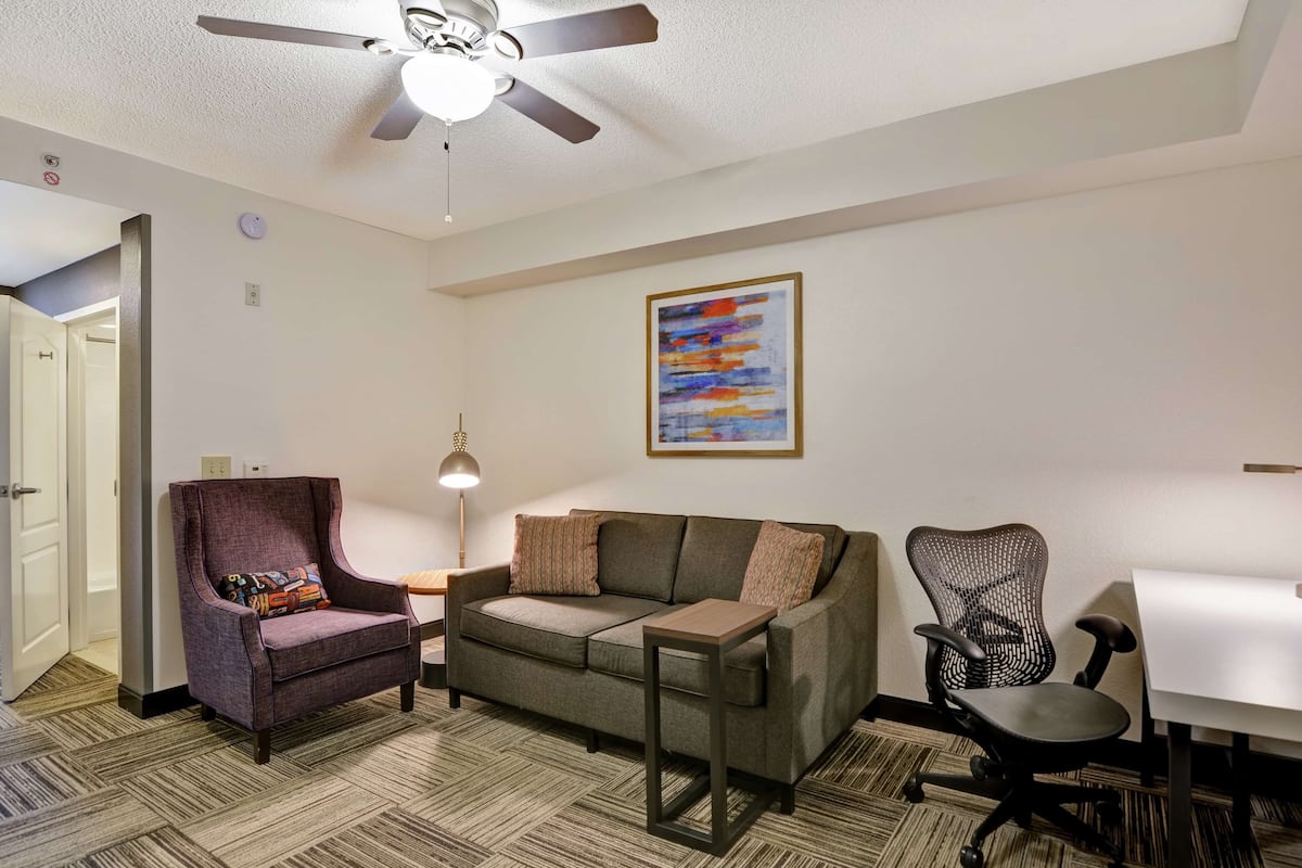 A modern living room with a sofa, armchair, and office chair, a ceiling fan, and a painting on the wall.