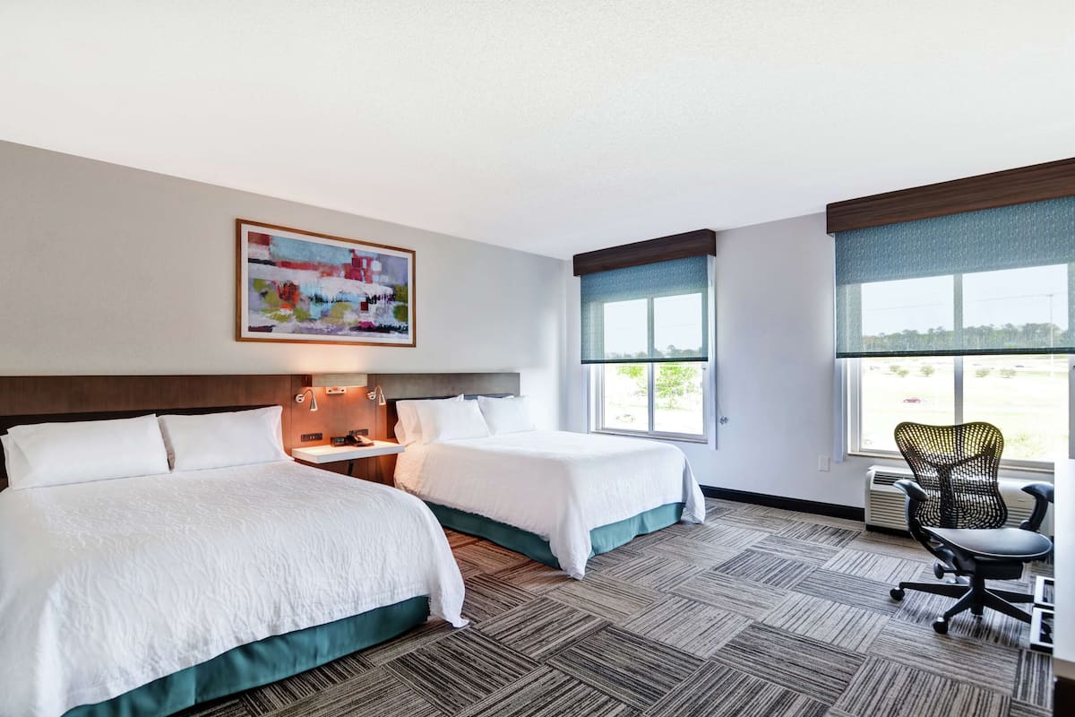 A hotel room with two beds, a desk, a chair, and a large window.