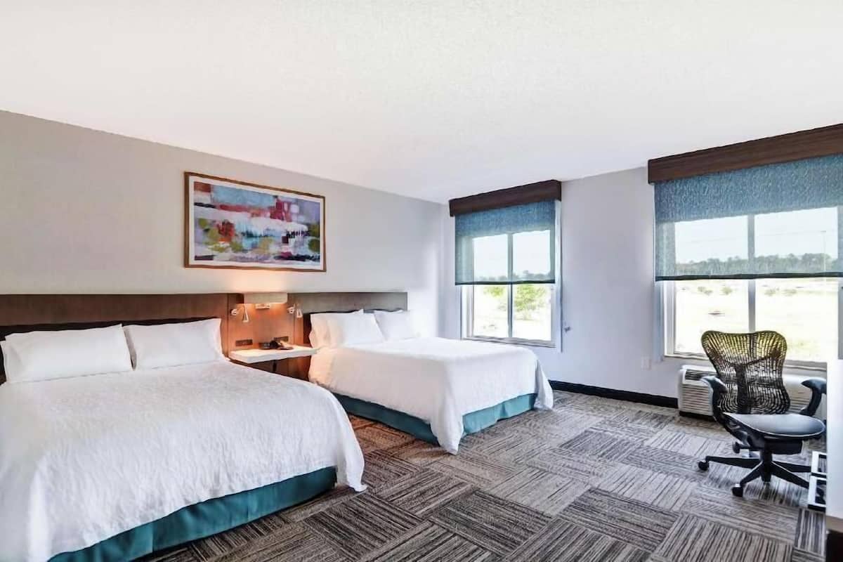 A hotel room with two beds, a desk, a chair, a painting, and large windows.
