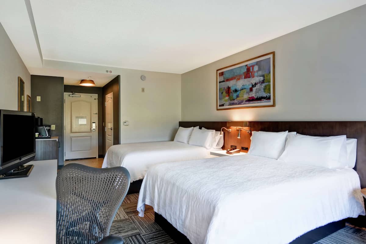 A hotel room with two beds, a desk with a television, a chair, and a painting on the wall.