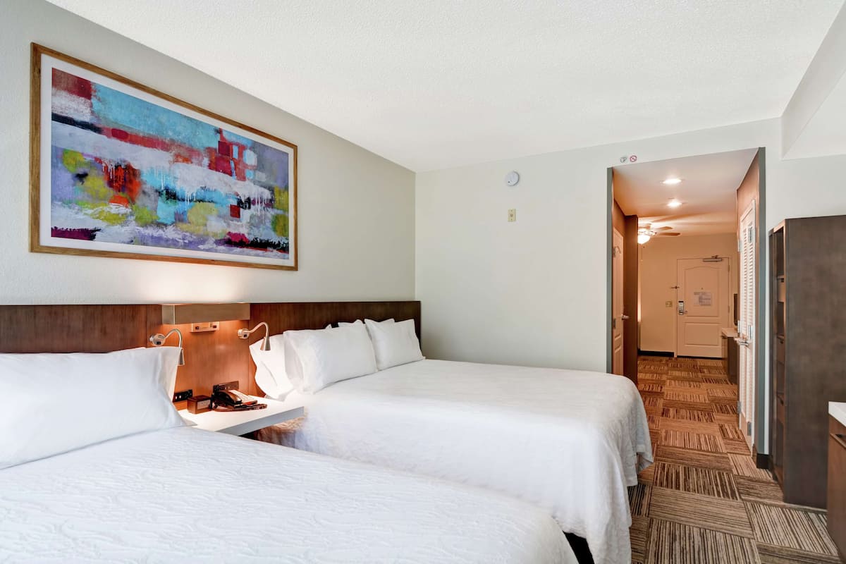 A hotel room with two beds, a painting on the wall, and a hallway leading to a door.
