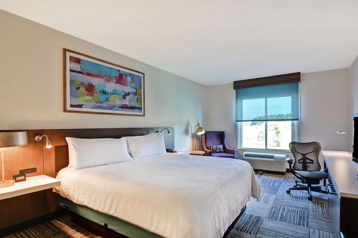 A hotel room with a large bed, a desk, a chair, a painting on the wall, and a window with a view.