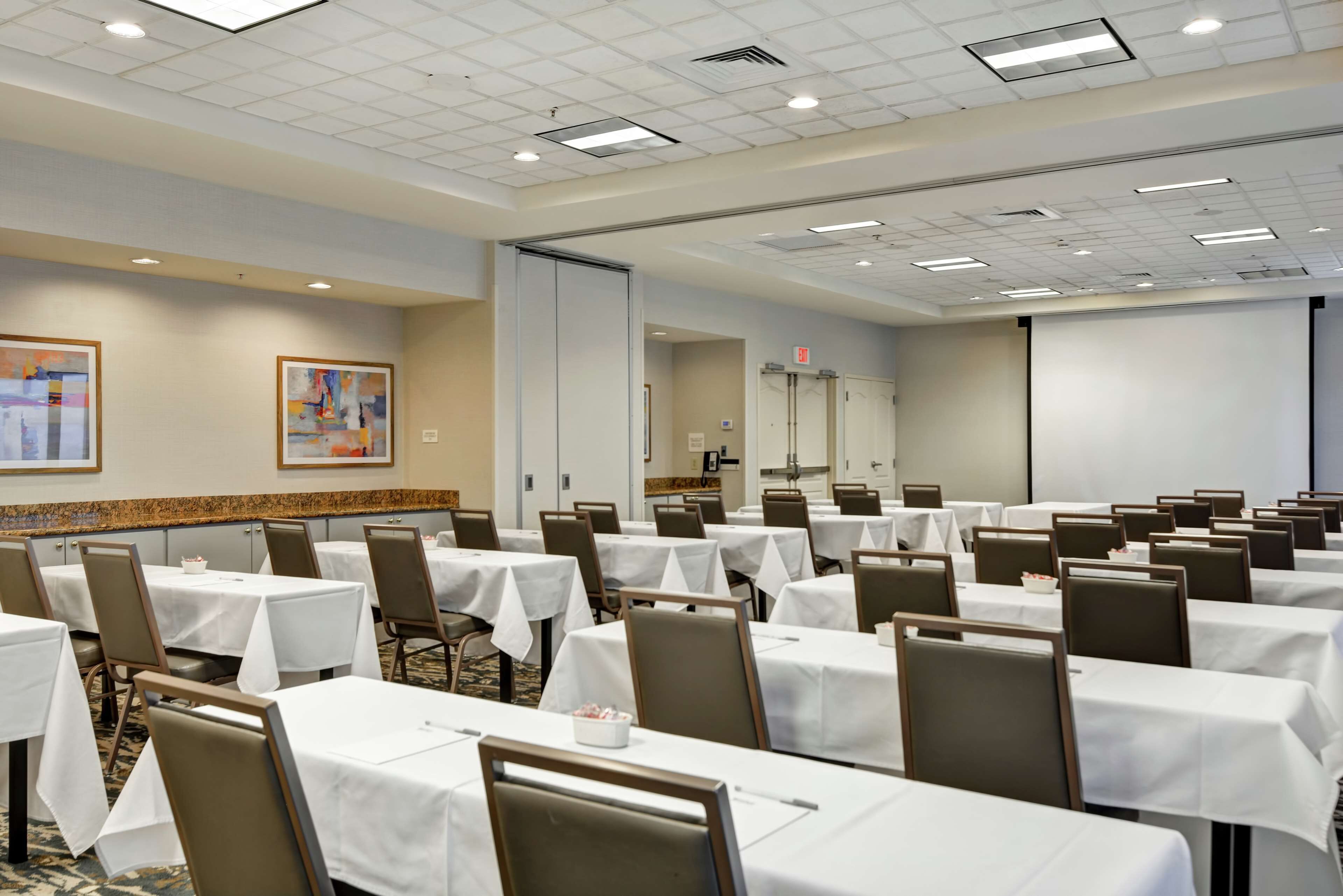 A conference room with multiple tables and chairs, a projector screen, and a door with an emergency exit sign.