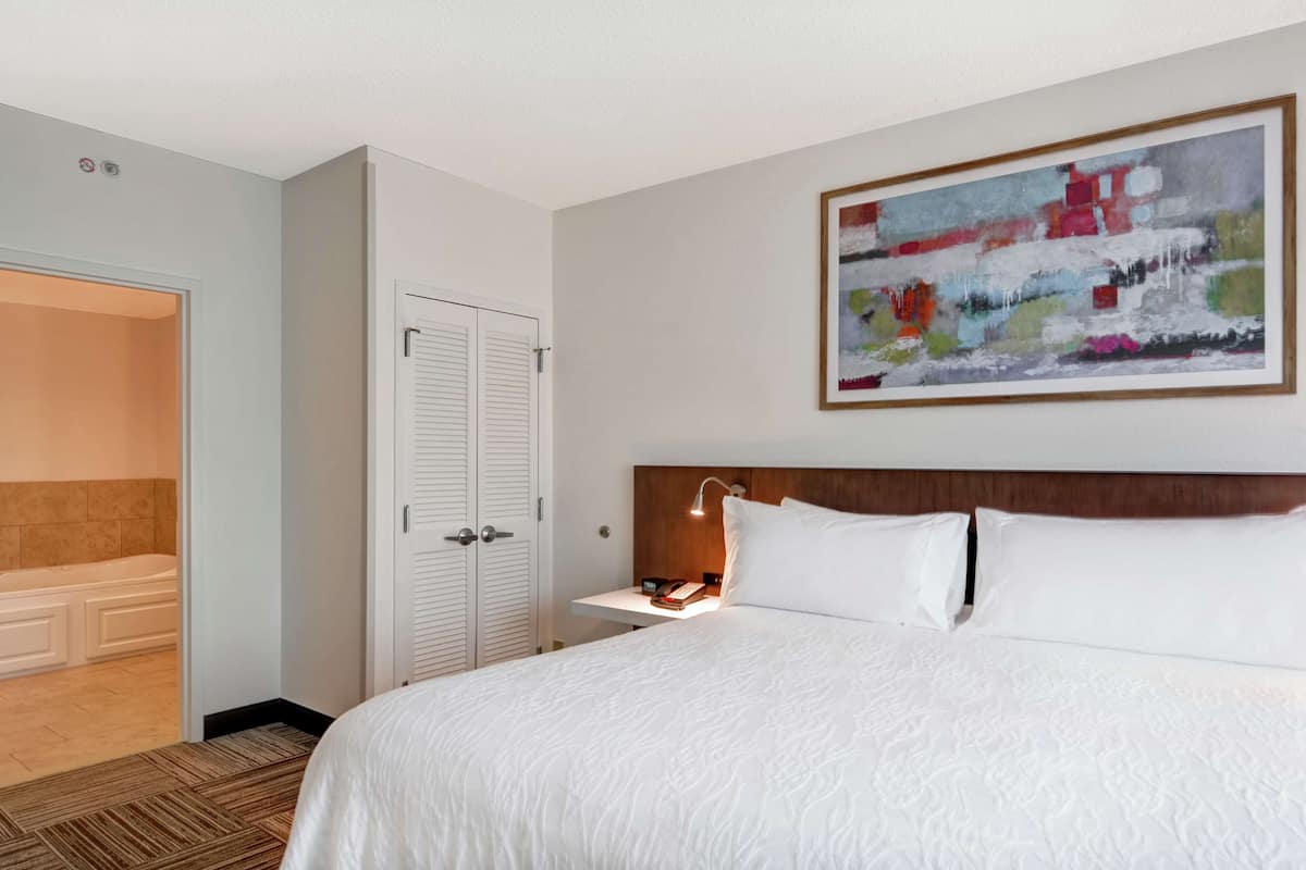A hotel room with a large bed, a bedside table with a lamp, a painting on the wall, and a bathroom with a bathtub.
