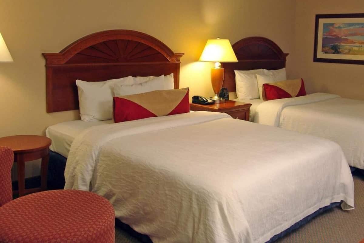A hotel room with two beds, a nightstand with a lamp, a chair, and a painting on the wall.