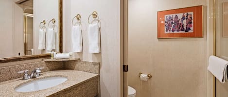 A bathroom with a granite countertop sink, a mirror, and a toilet. There are towels hanging on a towel rack, and a framed picture on the wall.