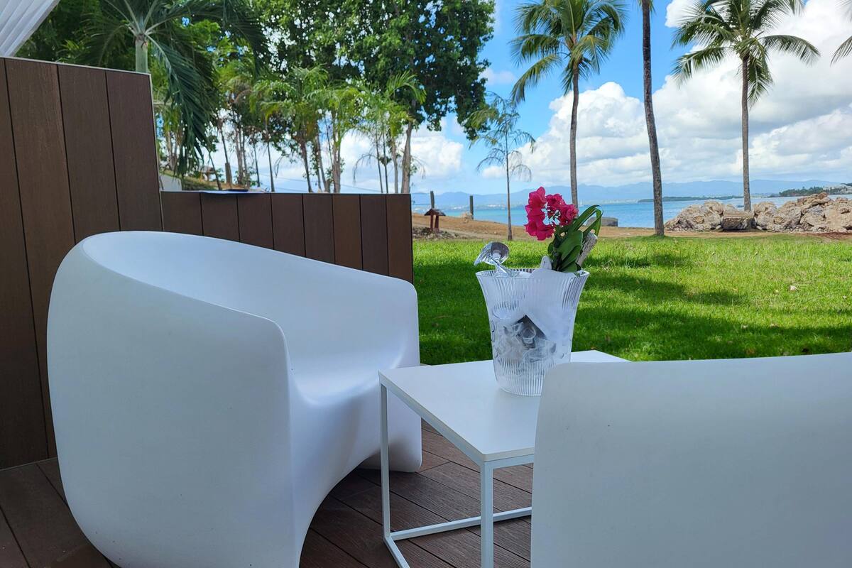 A modern outdoor seating area with a white chair and table, a vase with flowers, and a view of the ocean and palm trees.