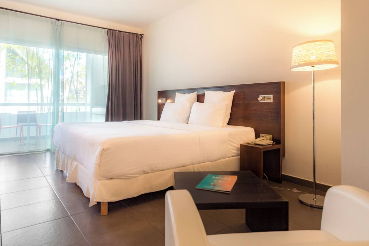 A hotel room with a large bed, a bedside table with a lamp, a small table with a book, and a chair.