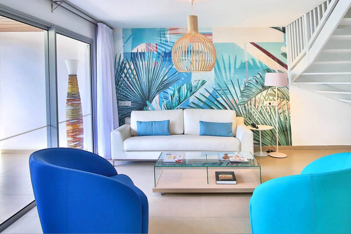 A modern living room with a white sofa, blue chairs, a glass coffee table, and a staircase.