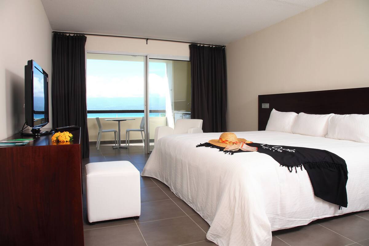 A hotel room with a large bed, a desk with a television, a balcony with a view of the sea, and a white chair.